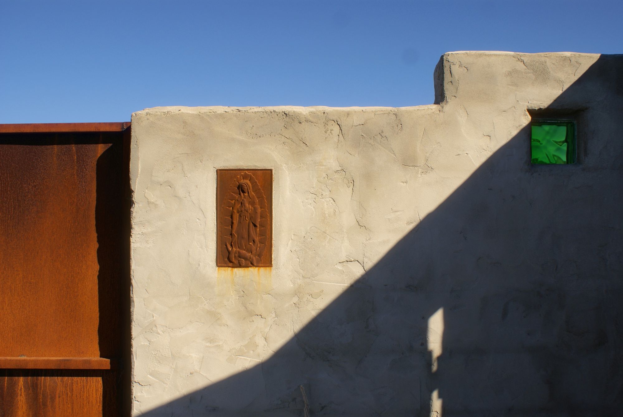 A relief of the Virgin of Guadalupe and a green glass block in the plaster wall of Las Sombras, beside the rusted gate